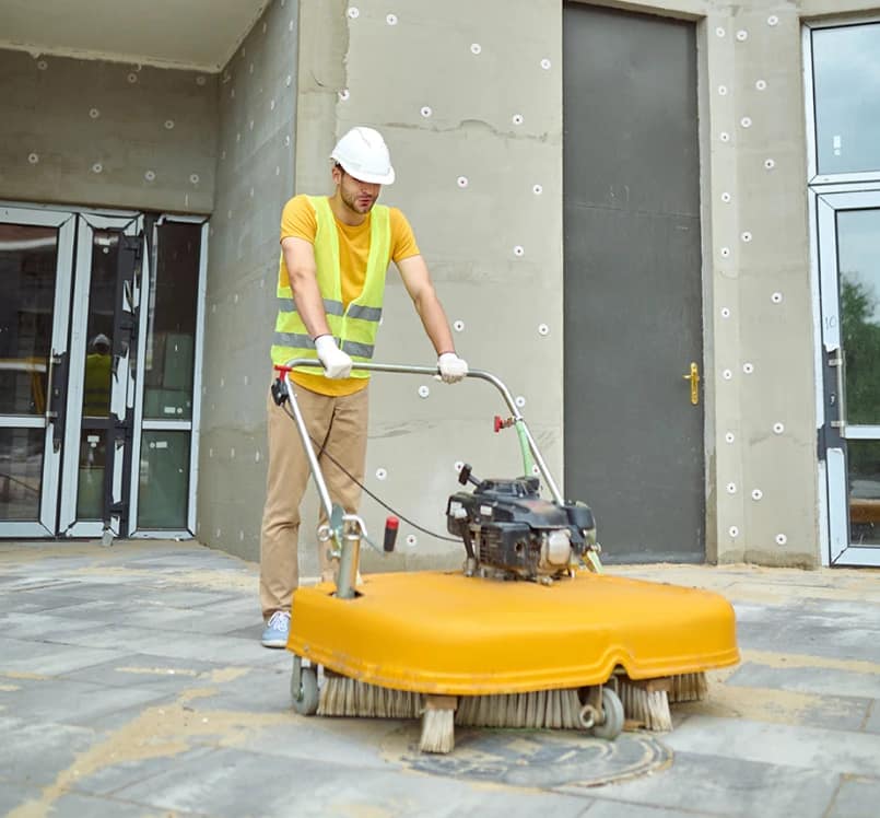 Construction Site Sweeper Worker in vest and hard hat using an outdoor floor sweeper on paving at a building site.